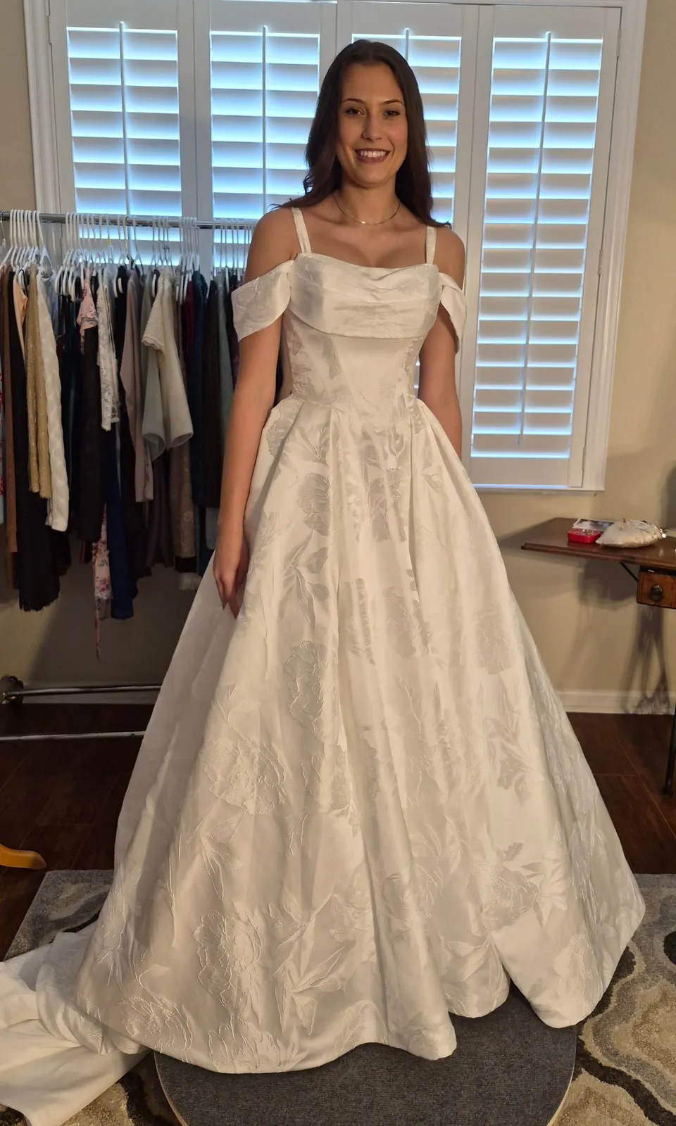 Smiling bride standing in a white floral jacquard ball gown with off-the-shoulder drape and thin straps